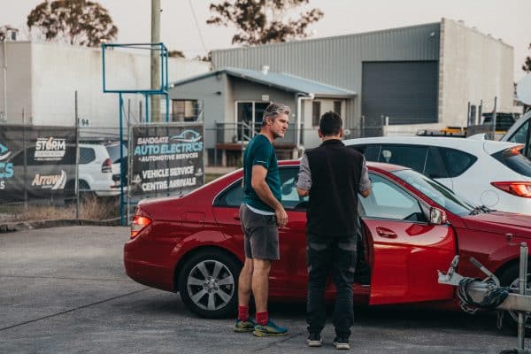 Two people discuss pre-purchase or safety checks in front of a red vehicle