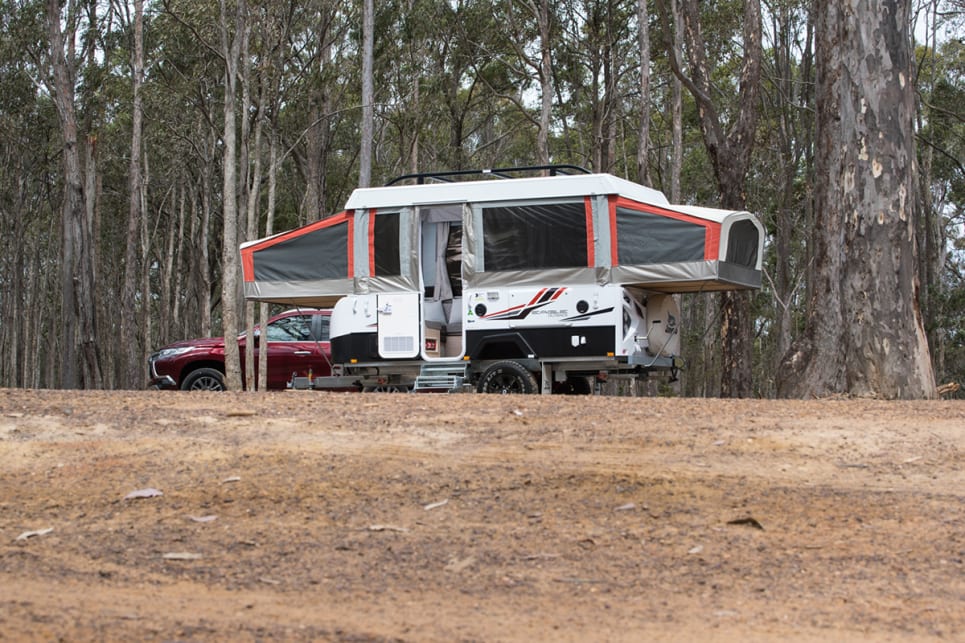 A small camper trailer with a pop-up top, parked next to a pickup truck in a wooded area