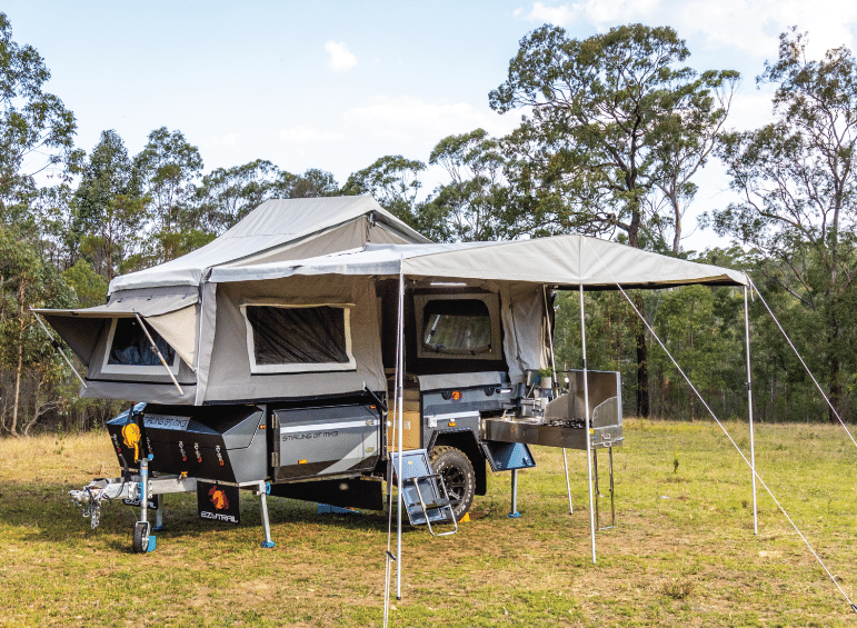 Vehicles parked on a dirt road in a forest clearing, with camping gear visible on the roof rack and chairs set up outside
